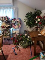 Wide view showing multiple faux floral arrangements in various containers including white ceramic and glass vases, red and white flowers, and leafy greenery. Several wreaths stacked on a chair and floor, partially wrapped in plastic.