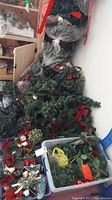 Assorted wrapped artificial Christmas trees, wreaths, bows, and garlands piled together and some stored in plastic bags.