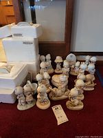 Overview of about sixteen Precious Moments figurines arranged on floor with some styrofoam packaging and wooden door background