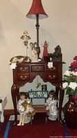 Wooden time out chair with porcelain dolls standing next to it, various angel figurines, resin hound dog, cranberry glass shoes, metal statue, brass bowl and table lamp on top of wooden furniture.