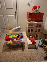 Wide shot showing wooden dollhouse, large container of colorful blocks, a small doll lying near dollhouse, and other toys and furniture arranged on carpet