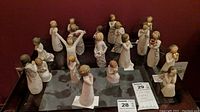 Full display of approximately 18 Willow Tree angel figurines with wire wings in varied poses on a mirrored surface against a dark red background.