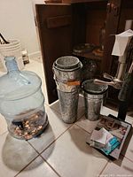 Full view of the large glass jug filled with coins positioned next to stacked galvanized buckets in front of wooden cabinet.