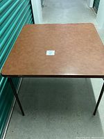 Photo of square collapsible bridge table with brown padded top and black metal legs, showing tabletop surface and some small marks.