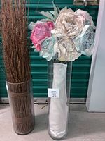 Two extra tall clear glass vases side by side. Left vase holds brown willow birch sticks bundled and tall. Right vase holds large pastel faux roses flowers in white, pink, blue hues with textured petals.