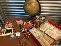Photo of lot showing vintage tabletop globe, vintage game boards including Chinese Checkers, cribbage board, various boxes including Jenga, Scrabble, electronic football, a Rubik's cube, View-Master, and shoebox with stamps.