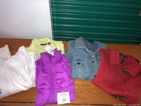 Lot of five ladies light jackets/tops displayed on a wooden table including red Ralph Lauren jacket, purple Adidas jacket, white Reebok jacket, light green Eddie Bauer jacket, and blue denim J Jill jacket.