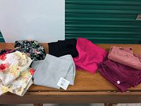 Nine pairs of ladies pants on a wooden table: floral patterns, solid black, pink, gray, peach, plum colors visible. Includes dress pants, cords, cotton chinos with floral prints.