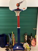 Photo shows adjustable Little Tikes basketball hoop with visible basketball graphic on the backboard, along with various sports equipment around the base including balls, helmets, rackets, and sports gear bags.