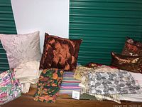 Four decorative pillows and folded table linens displayed on a table, showing variety in fabrics and patterns.