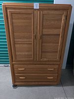 Front view of tall honey oak chest showing closed double doors with slatted design and two drawers below with wooden handles.