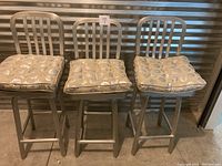 Three silver metal counter height swivel barstools with vertical slat backs shown side by side, each with removable beige cushions featuring circular patterns.