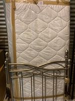 Photo showing wrapped twin mattress and metal headboard beneath it showing decorative metalwork and posts.