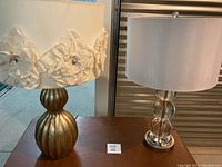 Two modern table lamps on wooden table with glass doors behind. One lamp has a gold ribbed base and cream shade with flower appliques. The other lamp has clear stacked glass spheres for base and plain white fabric shade.