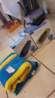 Photo showing the folded double sized beach towel with blue and yellow stripes, turquoise sand blanket, and three boxes (two showing pool loungers) on tiled floor.