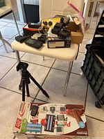 Overview photo showing various cameras, chargers, memory cards, and a tripod on a white chair and floor.