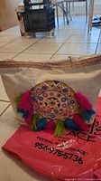 Front view of natural beige canvas tote bag with a colorful embroidered circular design in pink, orange, blue, and green, and attached colorful tassels.