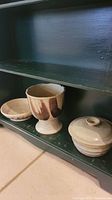 Three pottery items on green shelf: footed vase with chip, lidded covered pot, and shallow bowl