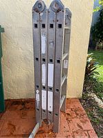 Folded aluminum industrial ladder leaning against wall showing side profile with rectangular cutouts and rust on hinge parts.
