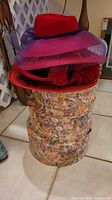 Four red hats stacked on top of three floral patterned vintage-style hat boxes. The hats include a red wool hat, a red hat with purple mesh brim and bow, and two red straw hats with decorative elements.