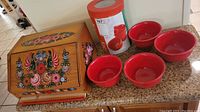 Front and side view of a wooden bread box decorated with colorful hand painted Eastern European floral designs, four red bowls placed nearby and a cutting board underneath the bread box.