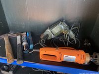 Photo showing electric drill, hammer with rubber grip, hammer with wooden handle, and cut-out tool on shelf.