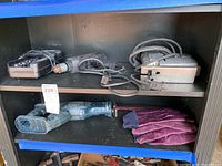 Photo showing the full set of power tools including the Black & Decker drill, two electric saws (reciprocating saw and belt sander), battery charger, battery box, and purple gloves all arranged on a black shelf.