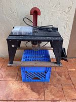 Black & Decker router mounted underneath a Vermont American Router/Saber Saw table on a blue crate, with a box of router bits on top.