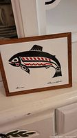 Framed ceramic tile with stylized salmon artwork by Clarence Wells on white ceramic.