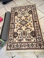 Full view of rectangular wool rug with detailed floral and geometric pattern, new fringe visible on edges, and new dark gray rug pad rolled beside it.