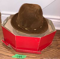 Brown velour fedora hat resting on red and white cardboard hat box base seen from side angle.