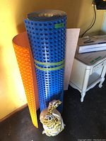 Photo showing two rolls of wide subfloor/vapor barrier mats, blue and orange, standing upright with visible texture of circular bumps.