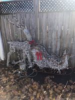 Photo of metal wire framed reindeer and sleigh decorated with white lights, placed outdoors with fallen leaves on the ground.