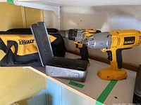 Dewalt cordless impact wrench, cordless drill, metal box of bits, and Dewalt carrying bag placed on a shelf.