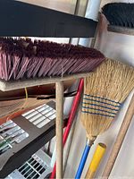 Close-up image of three push brooms showing black, purple synthetic, and natural straw bristles.