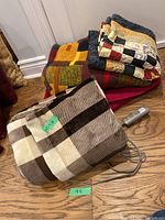 Full view of heated electric blanket with beige and brown checkered design next to quilted wool blankets in multicolored patchwork, red/gold pattern, and solid red colors.