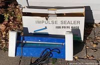 Blue impulse sealer with power cord next to original box and foam packaging shown outside on pavement.