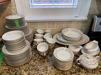 Photo showing full dish set including stacked plates, bowls, tea cups, and serving pieces on countertop under window.