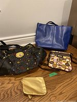 Lot of four assorted purses including blue tote, black designer style bag with colorful logos, brown and beige signature print small crossbody, and small gold pouch on wooden floor against white wall.