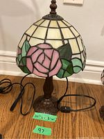 Full view of the stained glass lamp showing the pink rose and green leaf pattern on the shade, and visible black cord with inline switch.