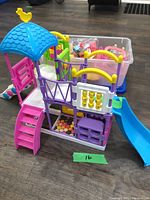 Front view of colorful plastic toy play set showing slide, stairs, roof with bird decoration, and lower interactive panels with accessories bin in background.