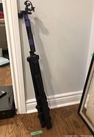 Folded black tripod with extendable legs, shown standing against a wall on a wooden floor.