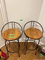 Two metal and wood bar stools showing black metal frames, circular wooden seats and curved backrests.