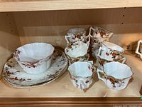 Photo showing several white porcelain tea cups and matching saucers with orange, brown, and black floral patterns and gold trim arranged on a wood shelf.