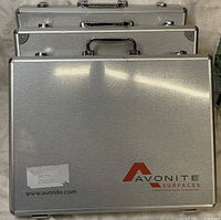 Three stacked rectangular aluminum carry cases with metal handles and framed edges, largest branded Avonite Surfaces on front