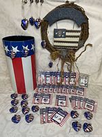Full lot display showing a grapevine wreath with 'Liberty' wooden flag plaque, a painted tin container with vertical red and white stripes and a blue band with white stars, filled with red, white, and blue heart-shaped glass ornaments, and numerous packages of patriotic body/face stickers spread out in front.