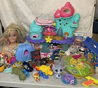Overall table view of assorted toys including dolls, play sets, plastic animals, and toy accessories.