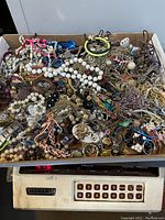 Overhead image of a box containing a large quantity of broken, vintage to modern jewelry pieces, beads, and findings, tangled and unorganized.
