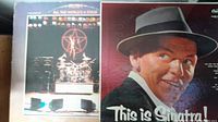 Photo of vintage vinyl records including Rush album 'All the World's a Stage' and Frank Sinatra album 'This is Sinatra!' showing wear and aging on sleeves.