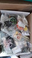 Photo shows multiple pairs of new earrings mixed in a cardboard box, still in packaging with visible variety of styles and colors.